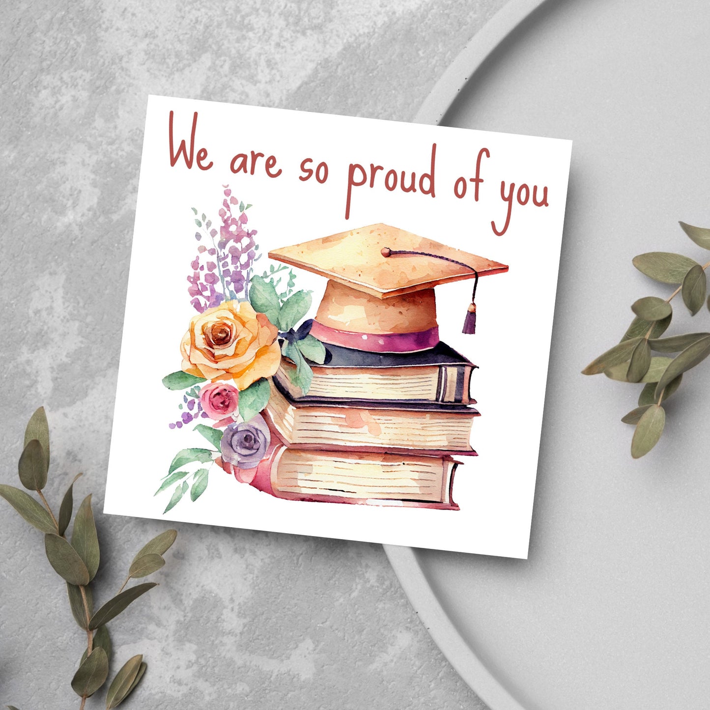 Image of a square graduation greeting card featuring a watercolour style image of a graduation cap on top of a stack of 3 books next to a floral arrangement. Above the image is the text 'we are so proud of you' in a handwritten font