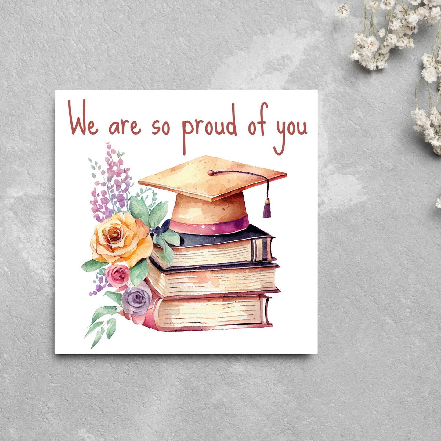 Image of a square graduation greeting card featuring a watercolour style image of a graduation cap on top of a stack of 3 books next to a floral arrangement. Above the image is the text 'we are so proud of you' in a handwritten font