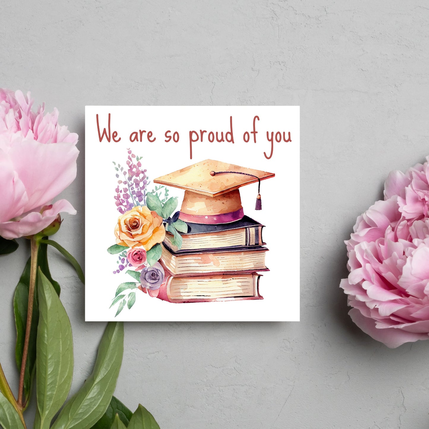 Image of a square graduation greeting card featuring a watercolour style image of a graduation cap on top of a stack of 3 books next to a floral arrangement. Above the image is the text 'we are so proud of you' in a handwritten font