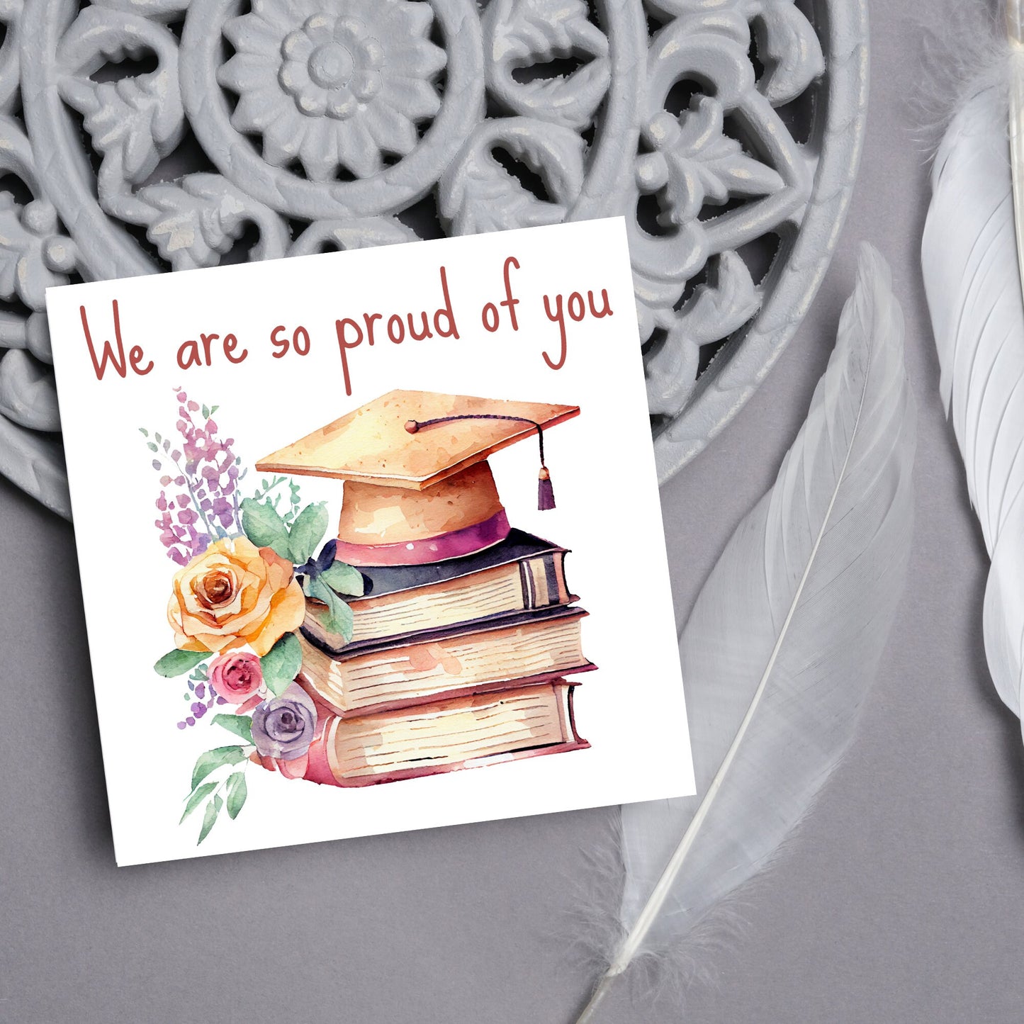 Image of a square graduation greeting card featuring a watercolour style image of a graduation cap on top of a stack of 3 books next to a floral arrangement. Above the image is the text 'we are so proud of you' in a handwritten font