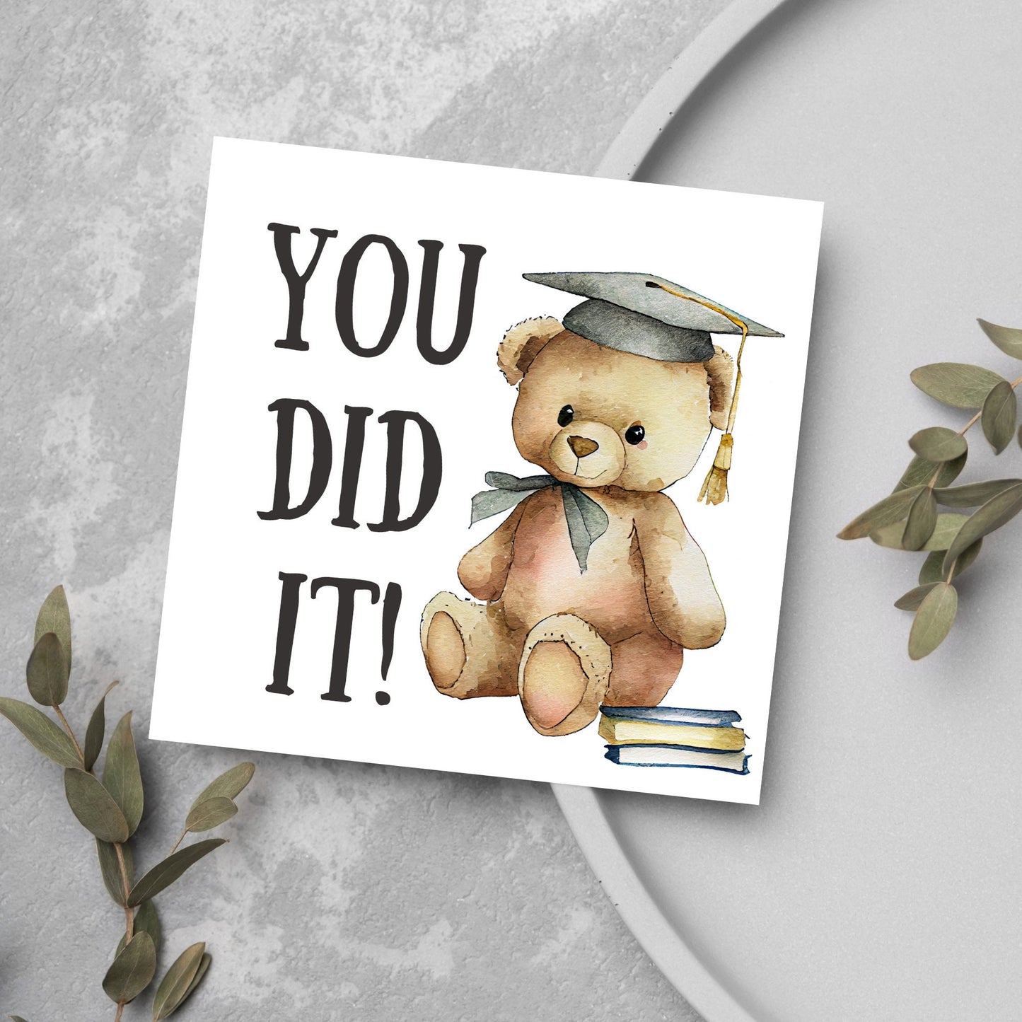 image of a square graduation greetings card featuring a watercolour illustration of a teddy bear wearing a graduation cap next to a pile of books. on the left of the bear is large black handwritten style text that reads 'you did it!'