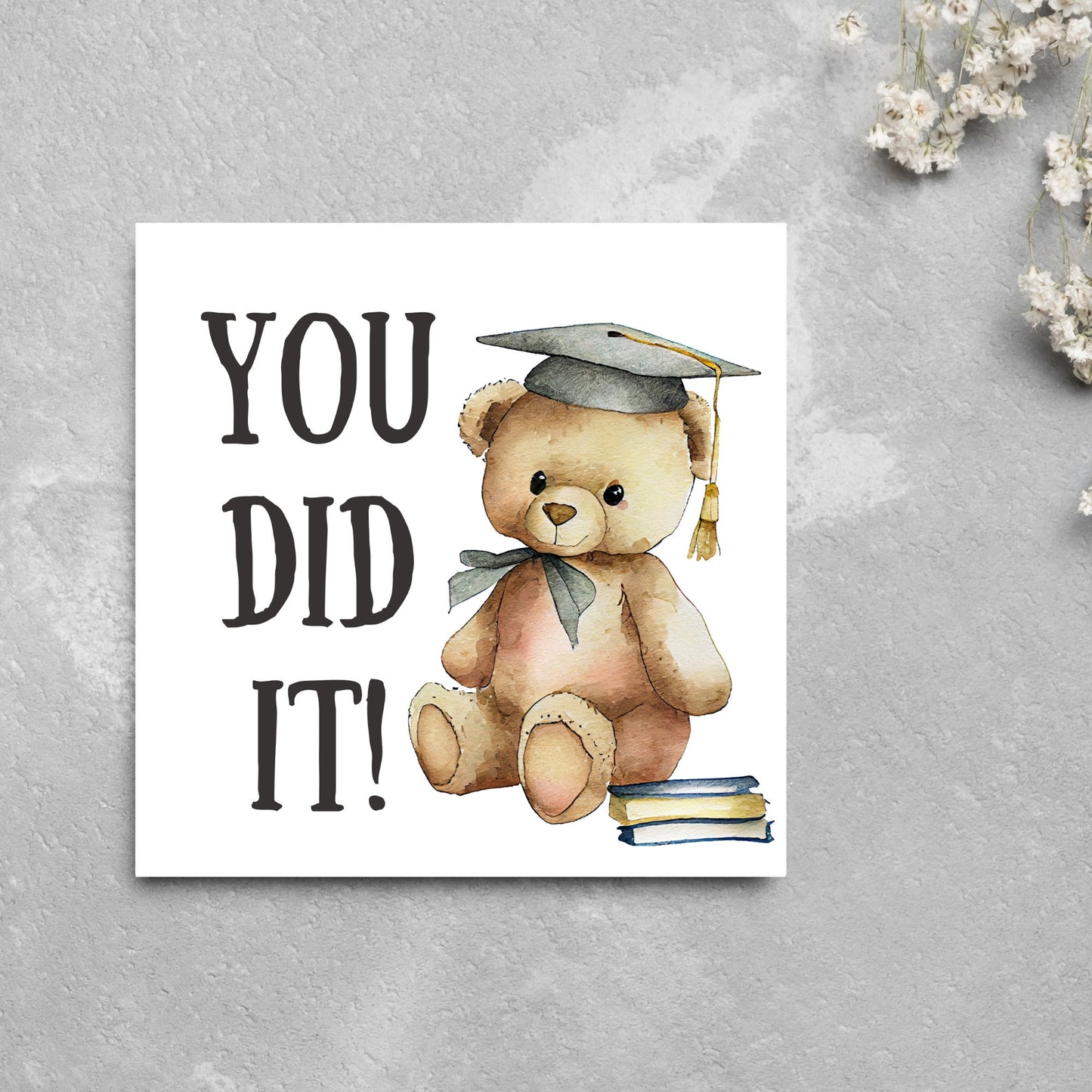 image of a square graduation greetings card featuring a watercolour illustration of a teddy bear wearing a graduation cap next to a pile of books. on the left of the bear is large black handwritten style text that reads 'you did it!'
