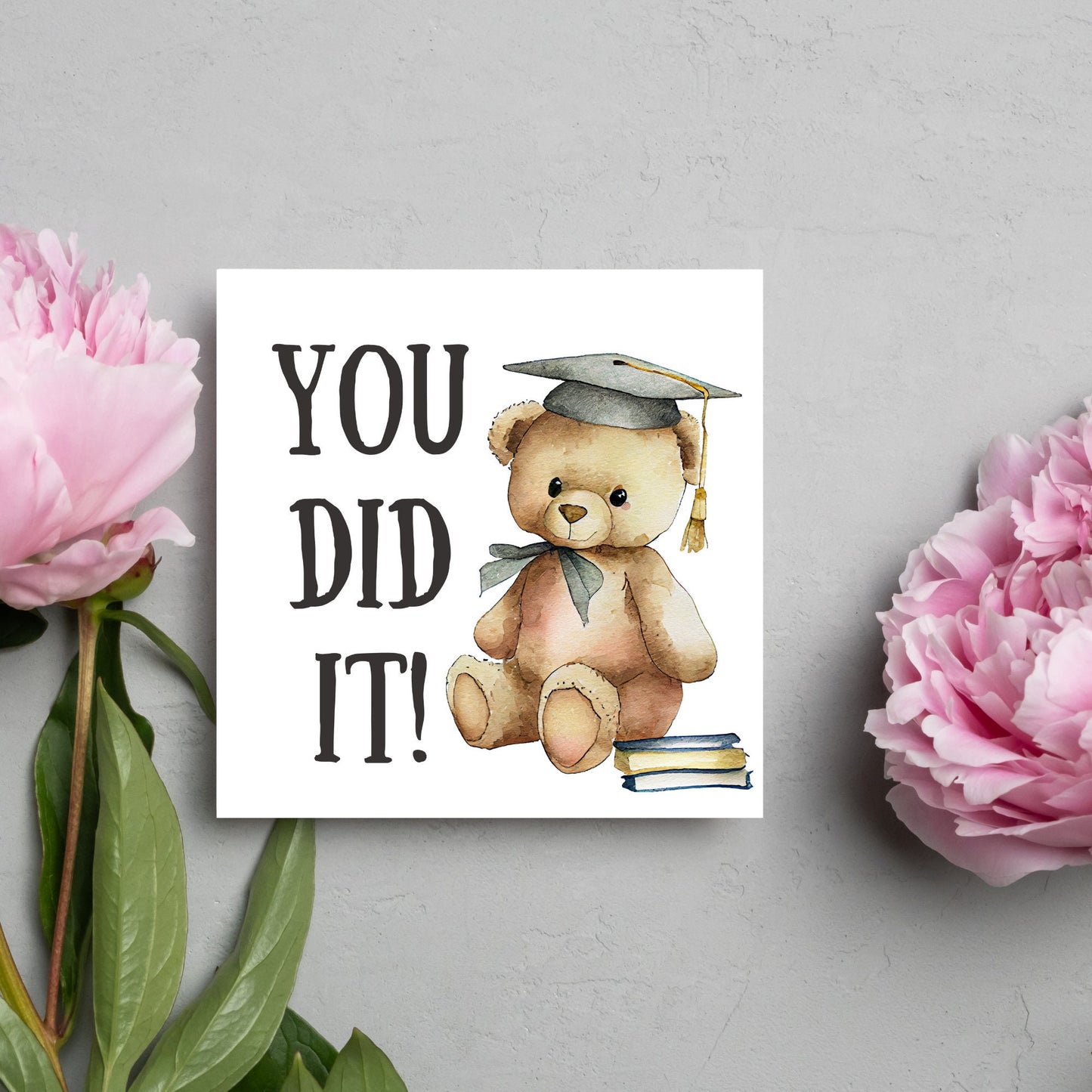 image of a square graduation greetings card featuring a watercolour illustration of a teddy bear wearing a graduation cap next to a pile of books. on the left of the bear is large black handwritten style text that reads 'you did it!'
