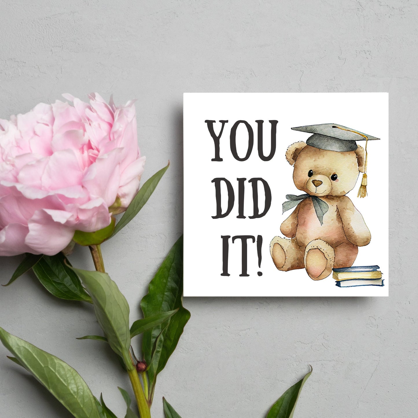 image of a square graduation greetings card featuring a watercolour illustration of a teddy bear wearing a graduation cap next to a pile of books. on the left of the bear is large black handwritten style text that reads 'you did it!'