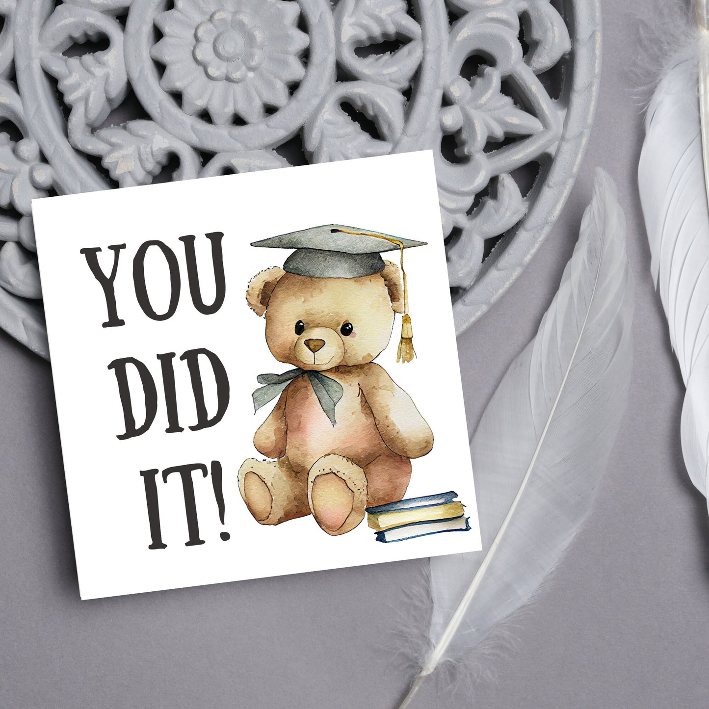 image of a square graduation greetings card featuring a watercolour illustration of a teddy bear wearing a graduation cap next to a pile of books. on the left of the bear is large black handwritten style text that reads 'you did it!'