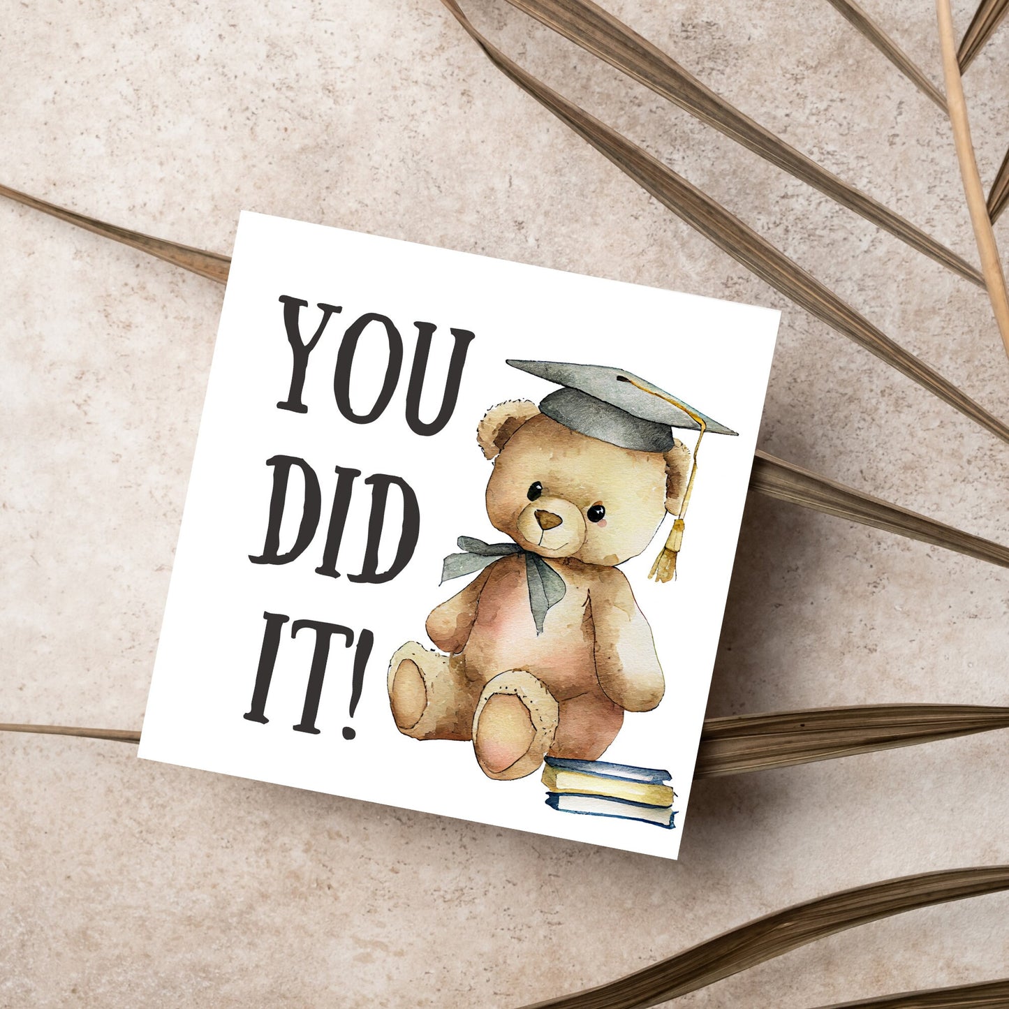 image of a square graduation greetings card featuring a watercolour illustration of a teddy bear wearing a graduation cap next to a pile of books. on the left of the bear is large black handwritten style text that reads 'you did it!'
