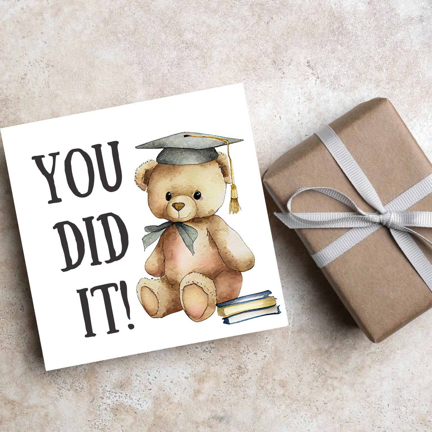 image of a square graduation greetings card featuring a watercolour illustration of a teddy bear wearing a graduation cap next to a pile of books. on the left of the bear is large black handwritten style text that reads 'you did it!'