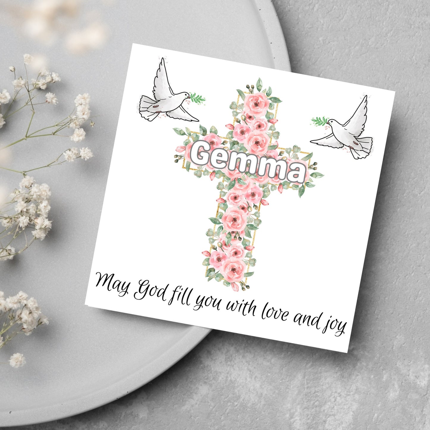 Square white card with a cross made of pink roses at the centre with the chosen name written across in white. either side of the cross is a white dove flying. At the bottom the text 'may god fill you with love and joy' is written