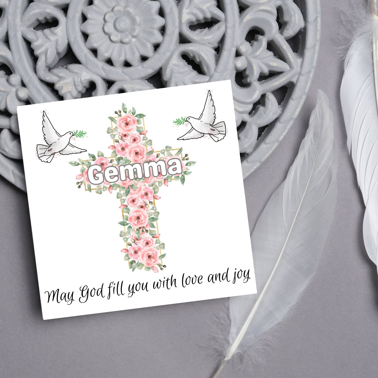 Square white card with a cross made of pink roses at the centre with the chosen name written across in white. either side of the cross is a white dove flying. At the bottom the text 'may god fill you with love and joy' is written