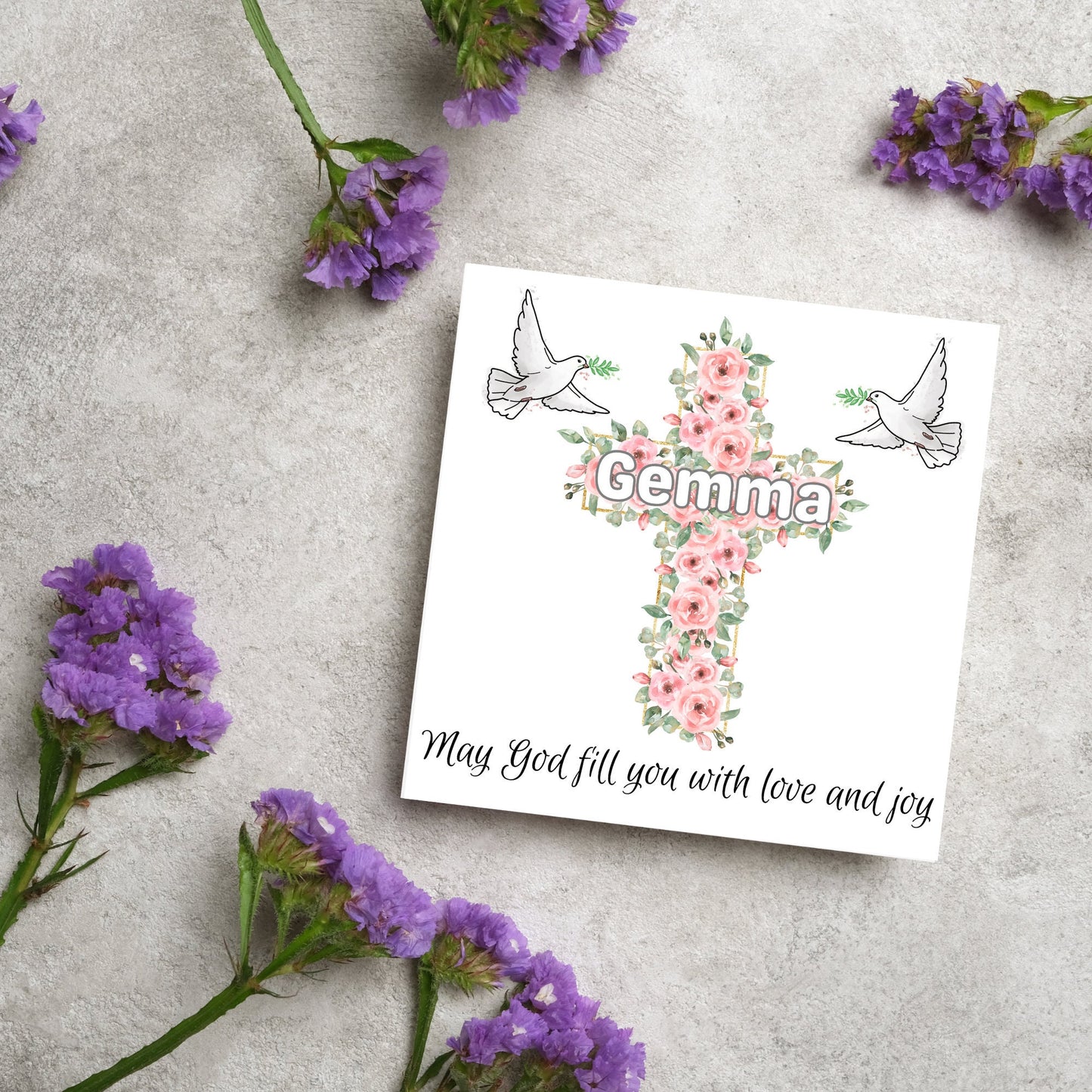 Square white card with a cross made of pink roses at the centre with the chosen name written across in white. either side of the cross is a white dove flying. At the bottom the text 'may god fill you with love and joy' is written