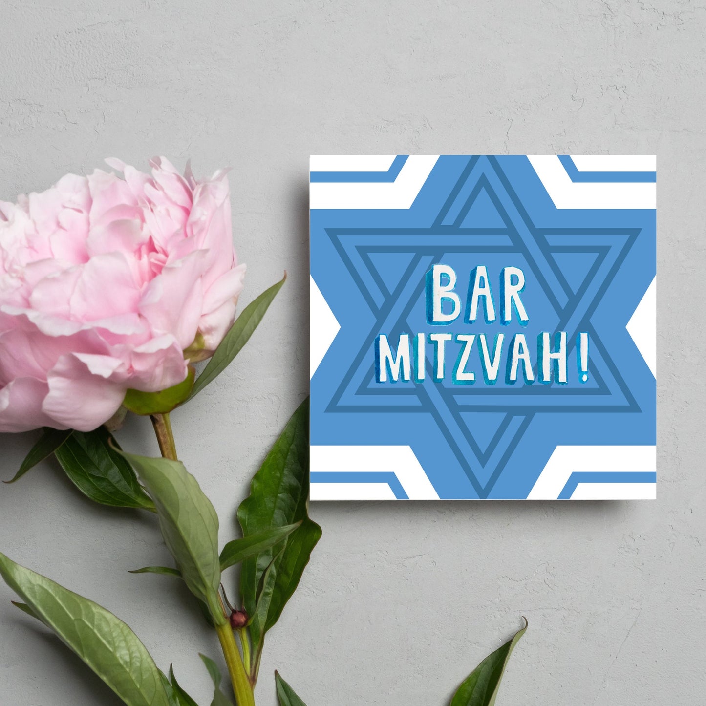 white and blue bar mitzvah card, geometric design with a repeating star of david at the centre in various shades of blue. At the centre of the card in white writing are the words 'bar mitzvah!' in capital letters in a handwritten style font