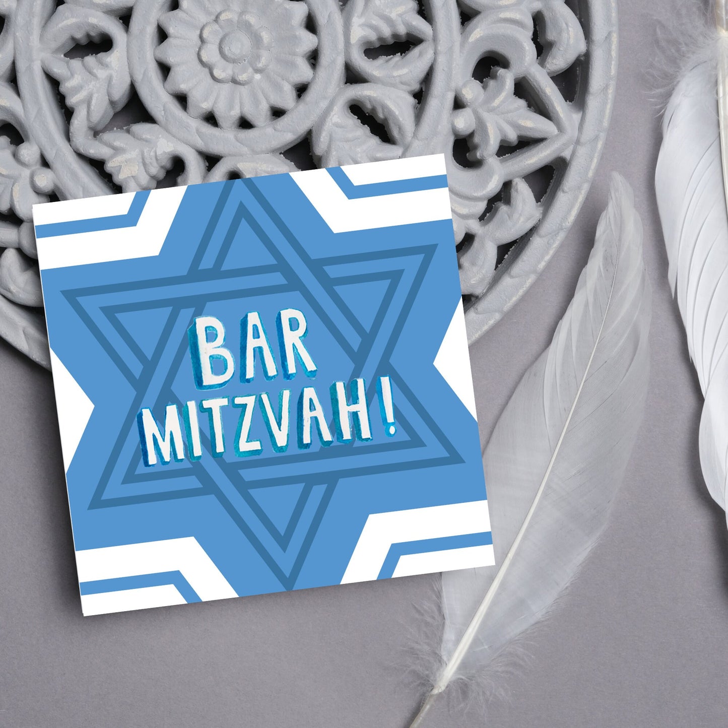white and blue bar mitzvah card, geometric design with a repeating star of david at the centre in various shades of blue. At the centre of the card in white writing are the words 'bar mitzvah!' in capital letters in a handwritten style font
