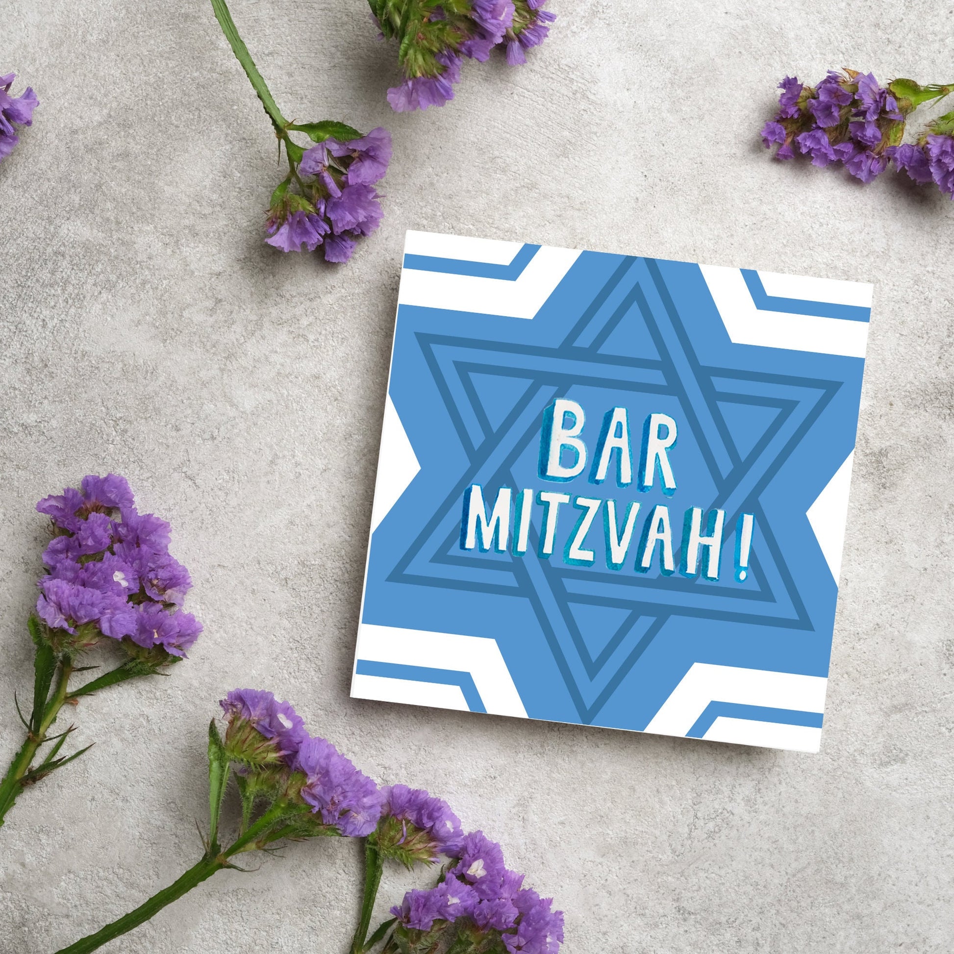 white and blue bar mitzvah card, geometric design with a repeating star of david at the centre in various shades of blue. At the centre of the card in white writing are the words 'bar mitzvah!' in capital letters in a handwritten style font