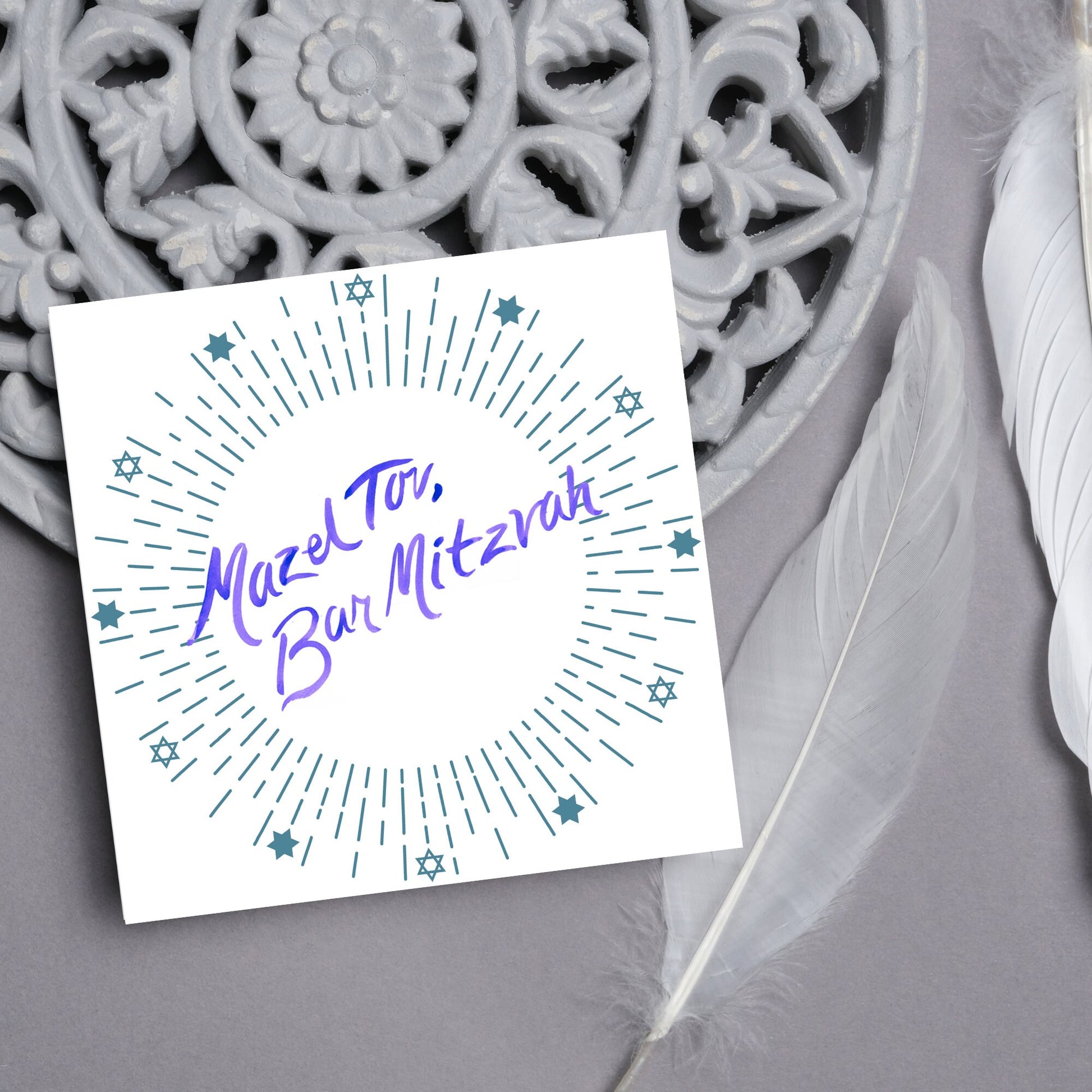 square bar mitzvah card with a white background and the text 'mazel tav, bar mitzvah' in the centre in a blue handwritten script style font. Surrounding the text is a circle made of concentric blue lines and 12 stars of david