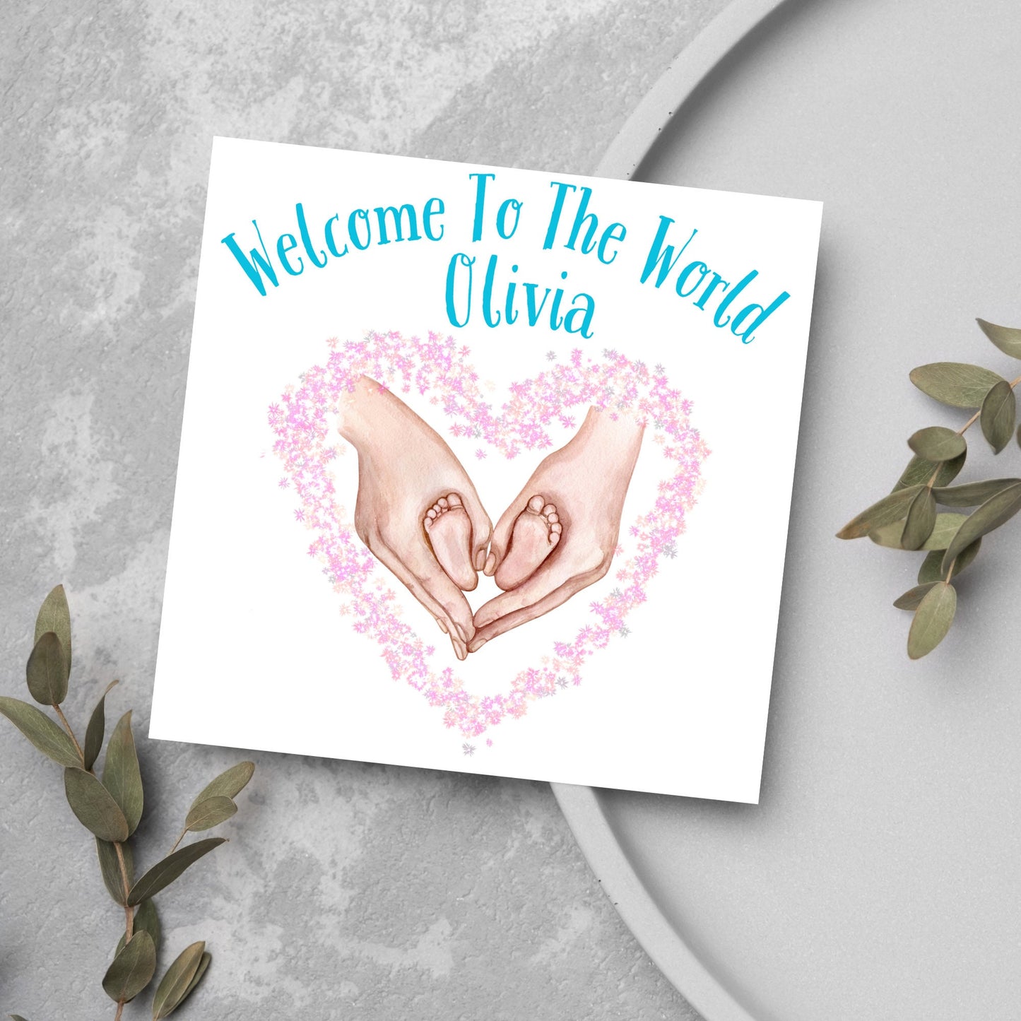 image of a square card with an illustration of hands holding newborn feet in a heart shape surrounded by pink flowers. Above the image is blue text that reads 'welcome to the world olivia' in a handwritten style font