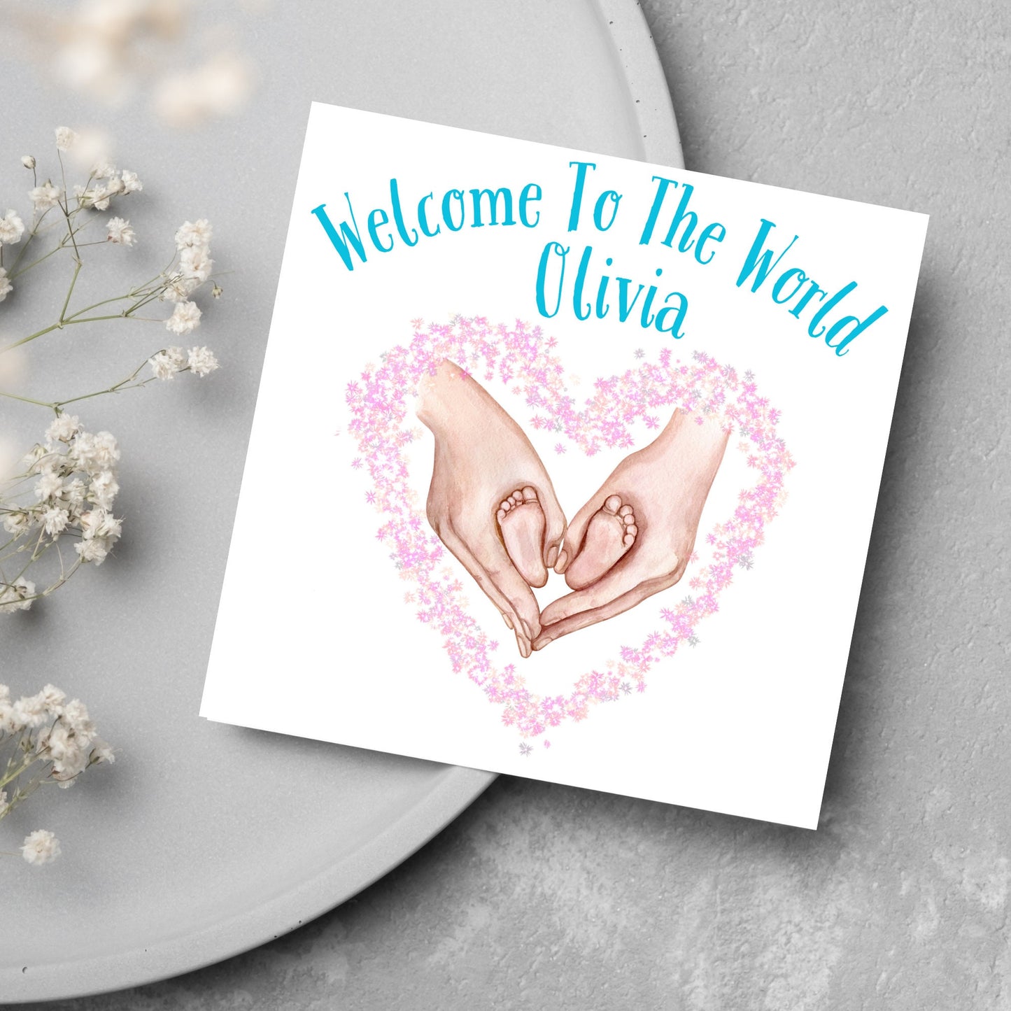 image of a square card with an illustration of hands holding newborn feet in a heart shape surrounded by pink flowers. Above the image is blue text that reads 'welcome to the world olivia' in a handwritten style font