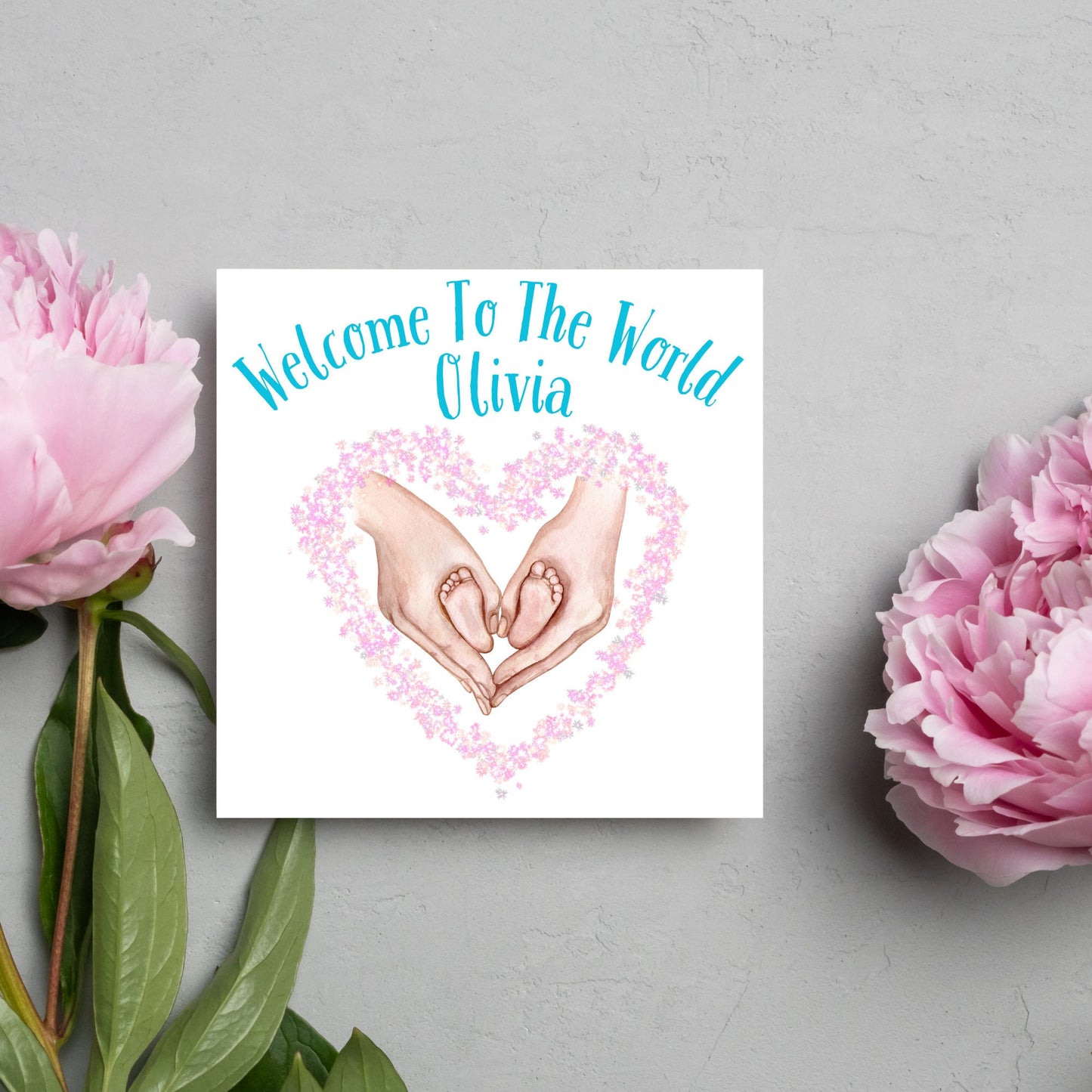 image of a square card with an illustration of hands holding newborn feet in a heart shape surrounded by pink flowers. Above the image is blue text that reads 'welcome to the world olivia' in a handwritten style font