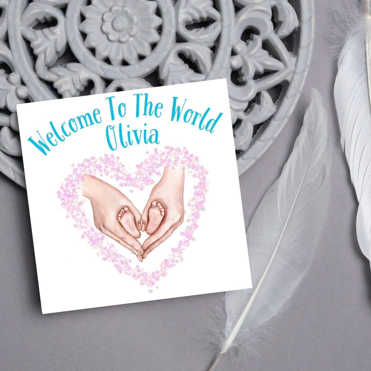 image of a square card with an illustration of hands holding newborn feet in a heart shape surrounded by pink flowers. Above the image is blue text that reads 'welcome to the world olivia' in a handwritten style font