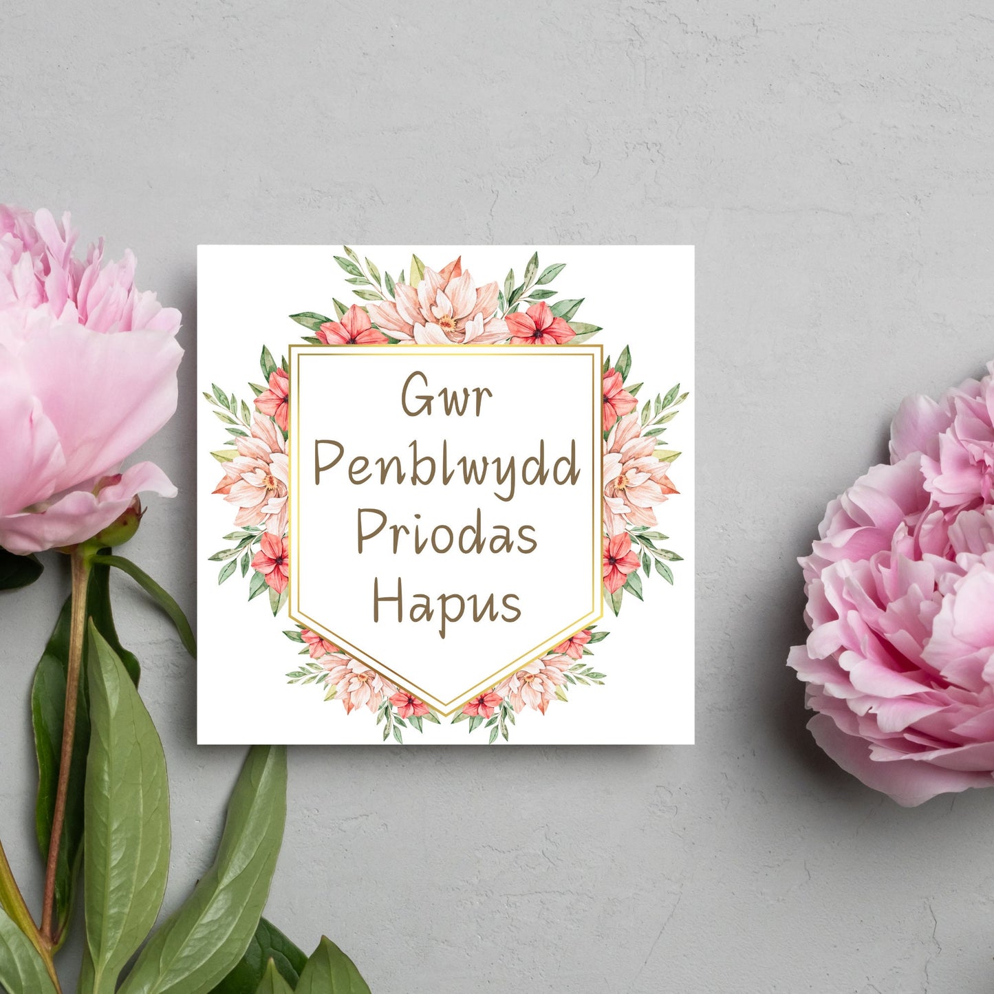 Image of a square wedding anniversary card with 'gwr penblwydd priodas hapus' in a subtle handwritten style font, which translates to 'happy wedding anniversary' in welsh. the text is against a white banner surrounded by pink flowers