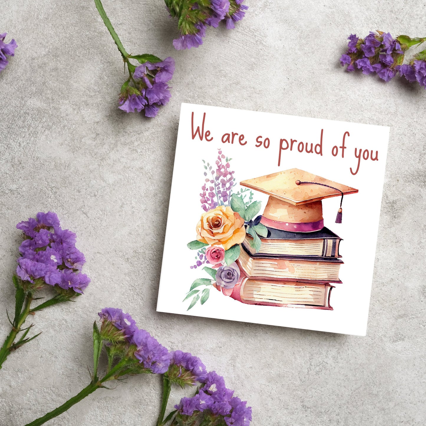 Image of a square graduation greeting card featuring a watercolour style image of a graduation cap on top of a stack of 3 books next to a floral arrangement. Above the image is the text 'we are so proud of you' in a handwritten font
