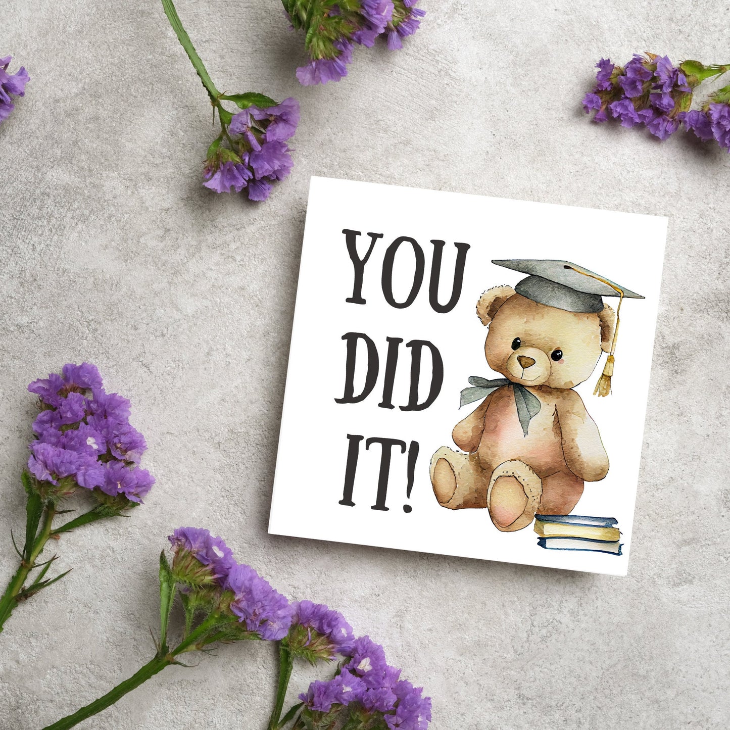 image of a square graduation greetings card featuring a watercolour illustration of a teddy bear wearing a graduation cap next to a pile of books. on the left of the bear is large black handwritten style text that reads 'you did it!'