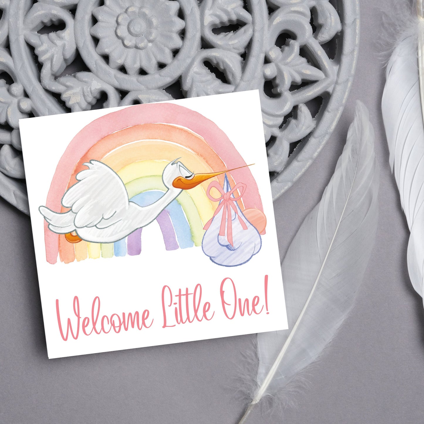 square new baby card with a flying cartoon stork delivering a swaddled baby against a watercolour rainbow in pastel shades. A cursive style text reads 'welcome little one' in a light pink font at the bottom of the card
