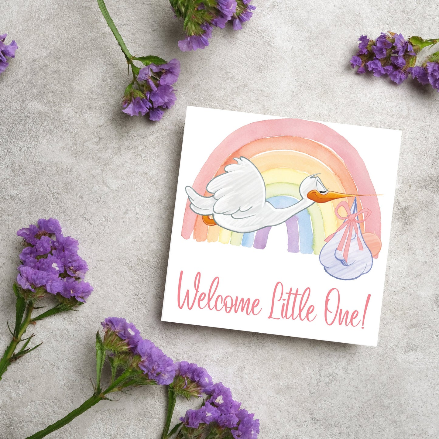 square new baby card with a flying cartoon stork delivering a swaddled baby against a watercolour rainbow in pastel shades. A cursive style text reads 'welcome little one' in a light pink font at the bottom of the card