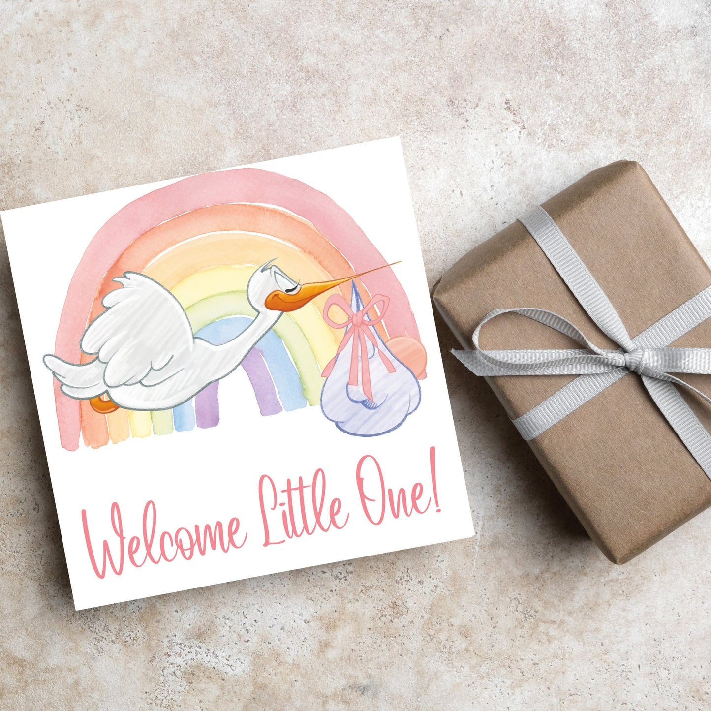 square new baby card with a flying cartoon stork delivering a swaddled baby against a watercolour rainbow in pastel shades. A cursive style text reads 'welcome little one' in a light pink font at the bottom of the card