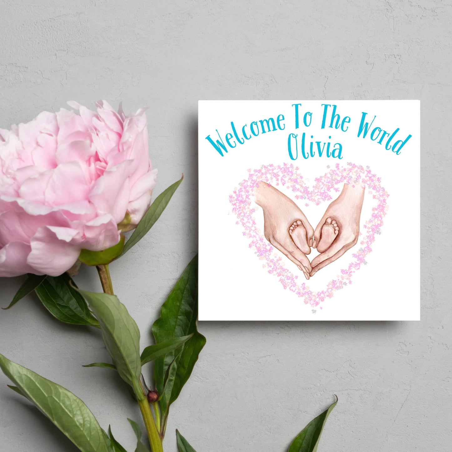 image of a square card with an illustration of hands holding newborn feet in a heart shape surrounded by pink flowers. Above the image is blue text that reads 'welcome to the world olivia' in a handwritten style font