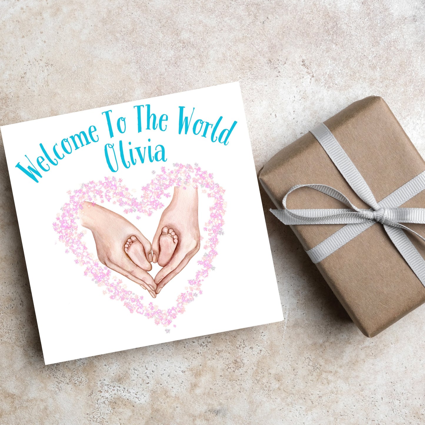image of a square card with an illustration of hands holding newborn feet in a heart shape surrounded by pink flowers. Above the image is blue text that reads 'welcome to the world olivia' in a handwritten style font