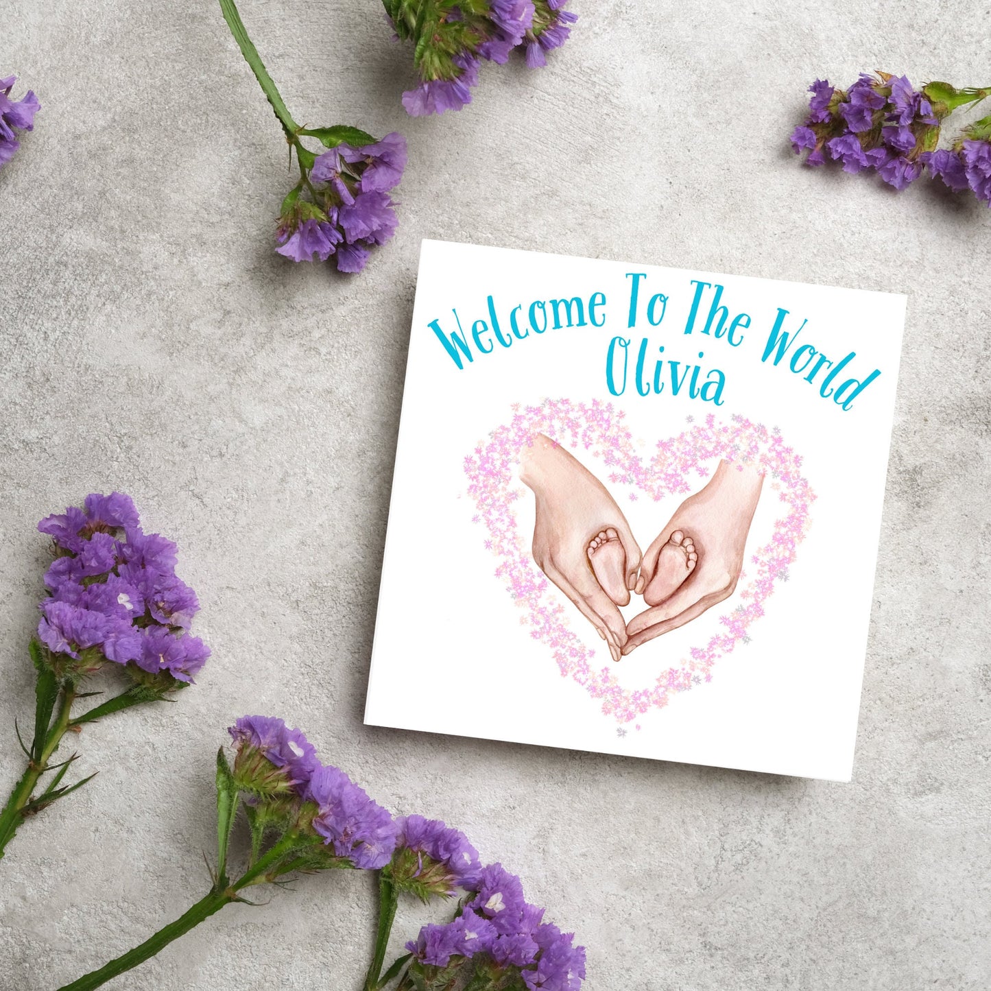 image of a square card with an illustration of hands holding newborn feet in a heart shape surrounded by pink flowers. Above the image is blue text that reads 'welcome to the world olivia' in a handwritten style font