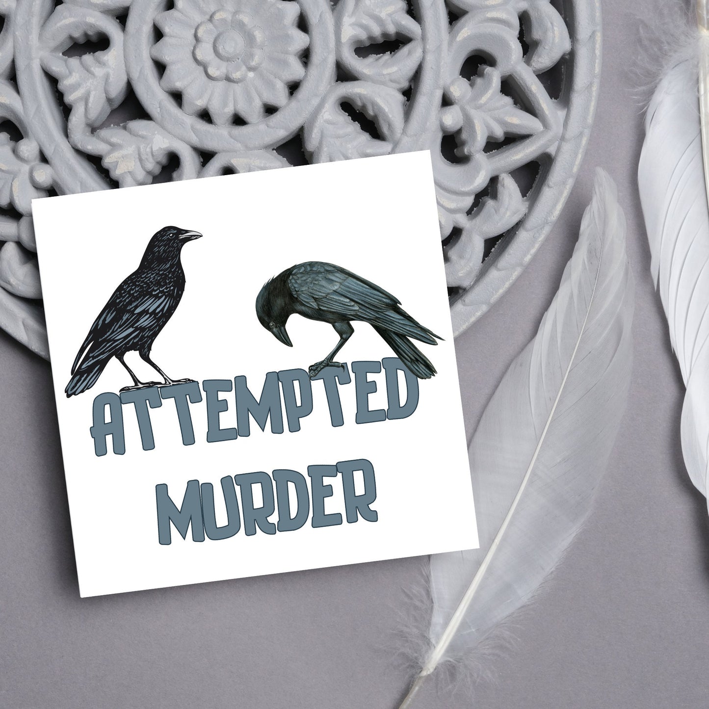 square card with the words attempted murder and 2 crows sitting on top of the text (a murder is the collective name 3 or more crows)