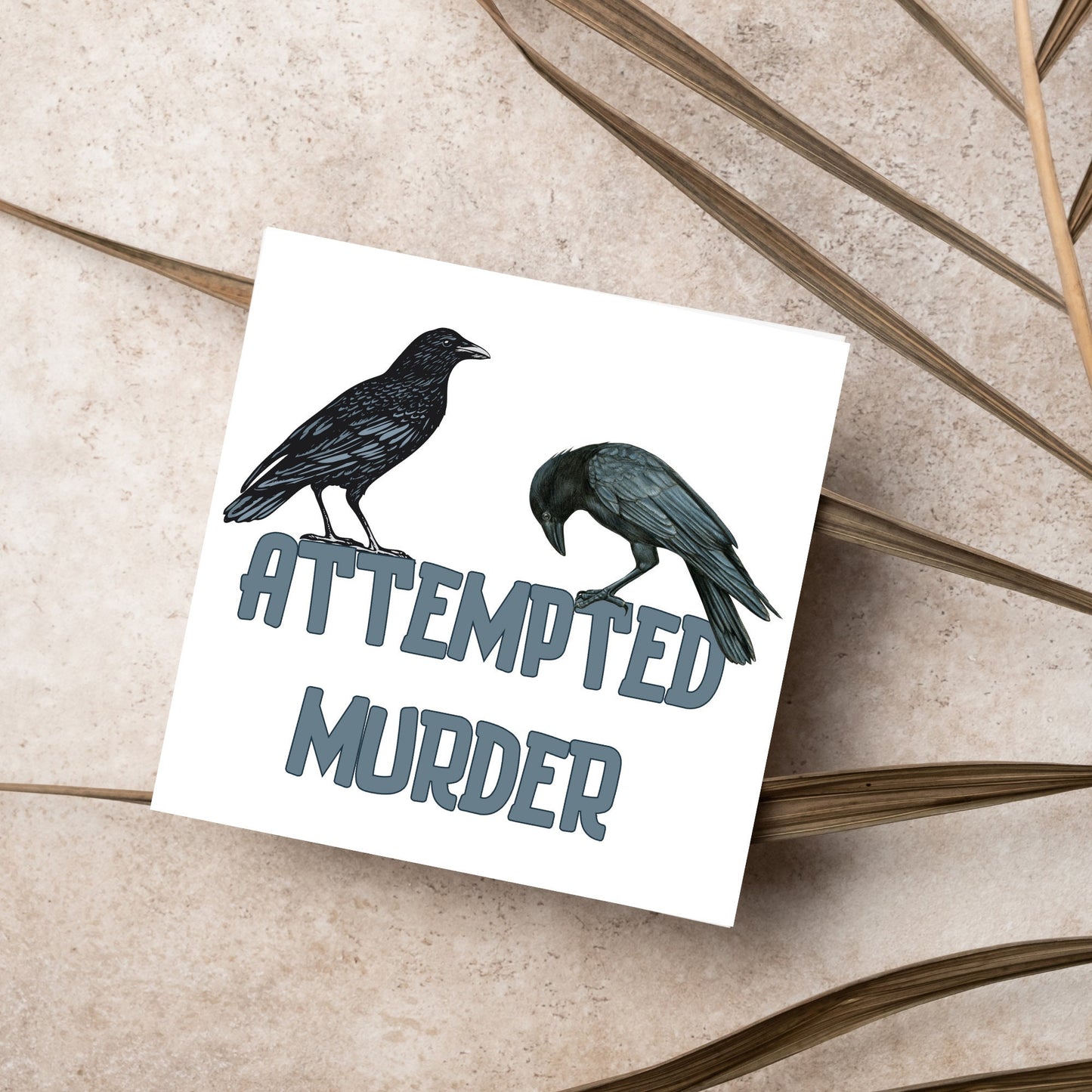 square card with the words attempted murder and 2 crows sitting on top of the text (a murder is the collective name 3 or more crows)