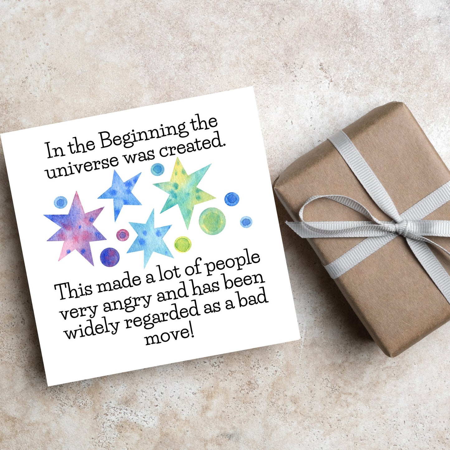 square card with watercolour abstract stars and planets and the text - in the beginning the universe was created. This made a lot of people very angry and has been widely regarded as a bad move! from the hitchhikers guide to the galaxy