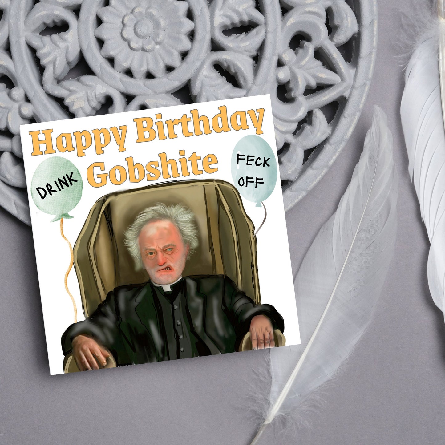 square birthday card with a hand drawn image of father jack from the tv show father ted. he is sitting in his chair looking mean with the text happy birthday gobshite above him. 2 balloons are next to him with feck off and drink on them