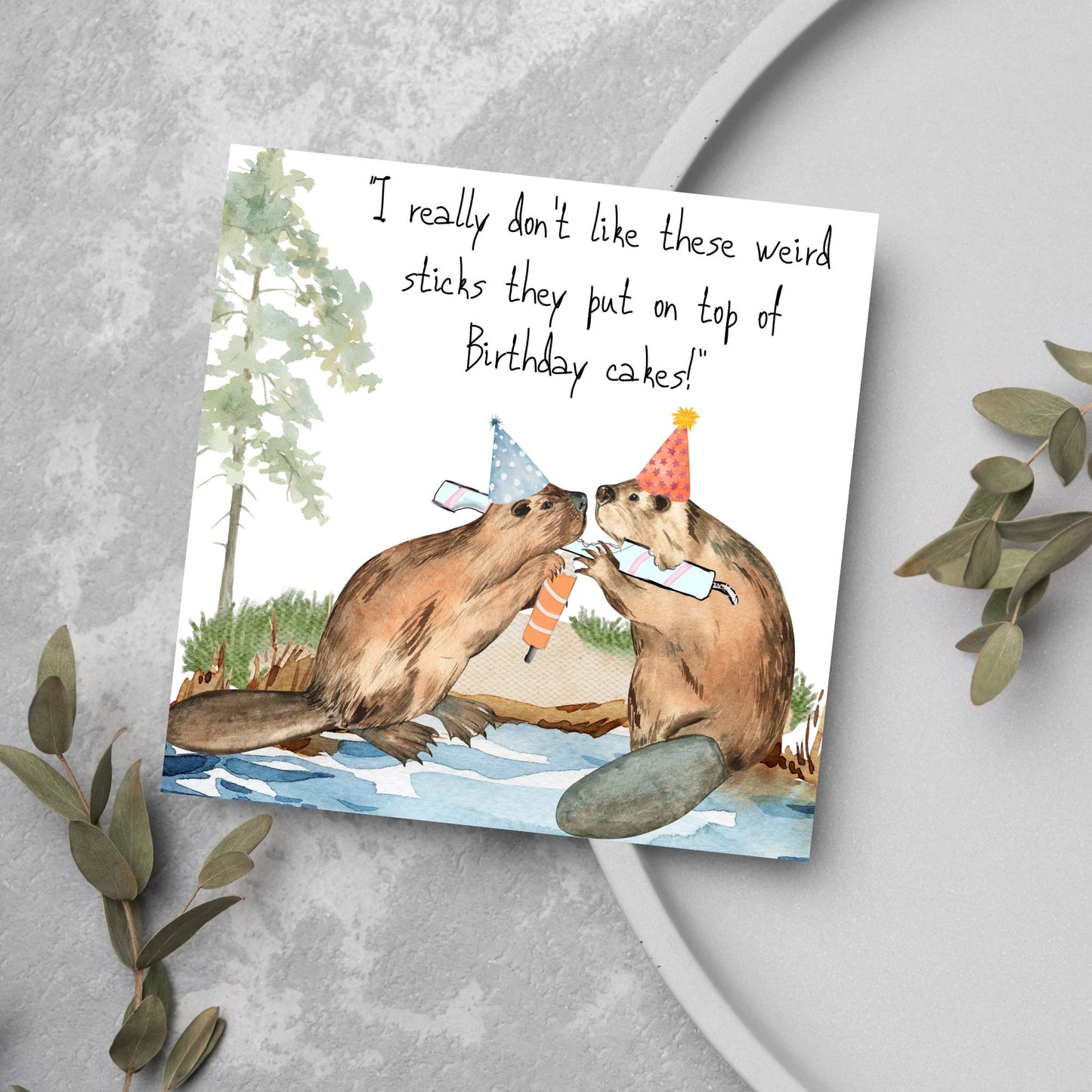 Image of a square birthday card with a watercolour image of 2 beavers with party hats on, chewing candles and handwritten style text above them that reads - i really don't like these weird sticks they put on top of birthday cakes!