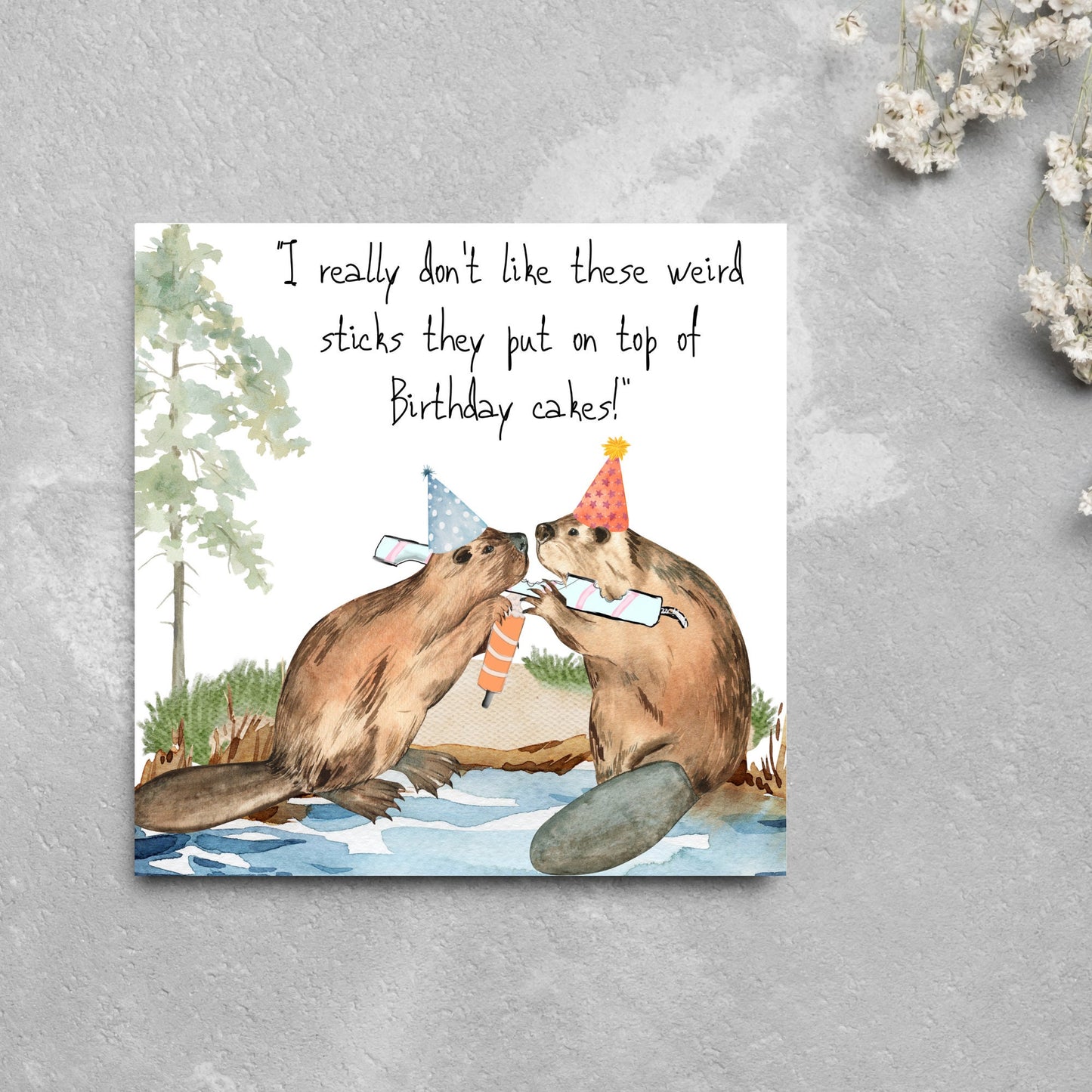 Image of a square birthday card with a watercolour image of 2 beavers with party hats on, chewing candles and handwritten style text above them that reads - i really don't like these weird sticks they put on top of birthday cakes!