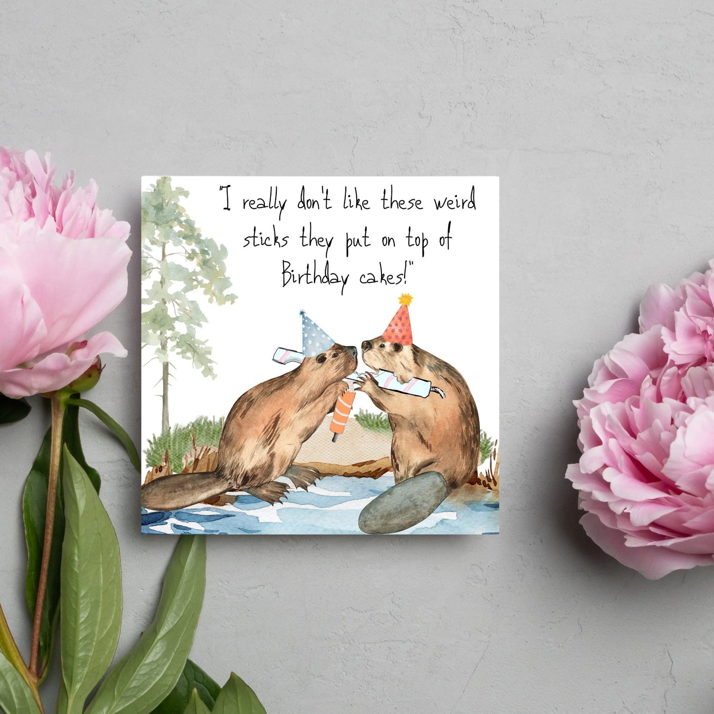 Image of a square birthday card with a watercolour image of 2 beavers with party hats on, chewing candles and handwritten style text above them that reads - i really don't like these weird sticks they put on top of birthday cakes!