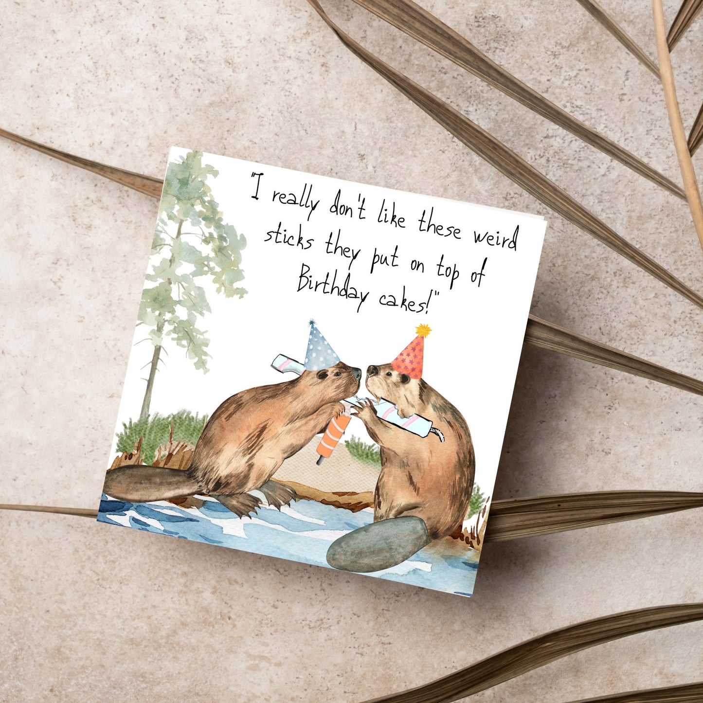 Image of a square birthday card with a watercolour image of 2 beavers with party hats on, chewing candles and handwritten style text above them that reads - i really don't like these weird sticks they put on top of birthday cakes!