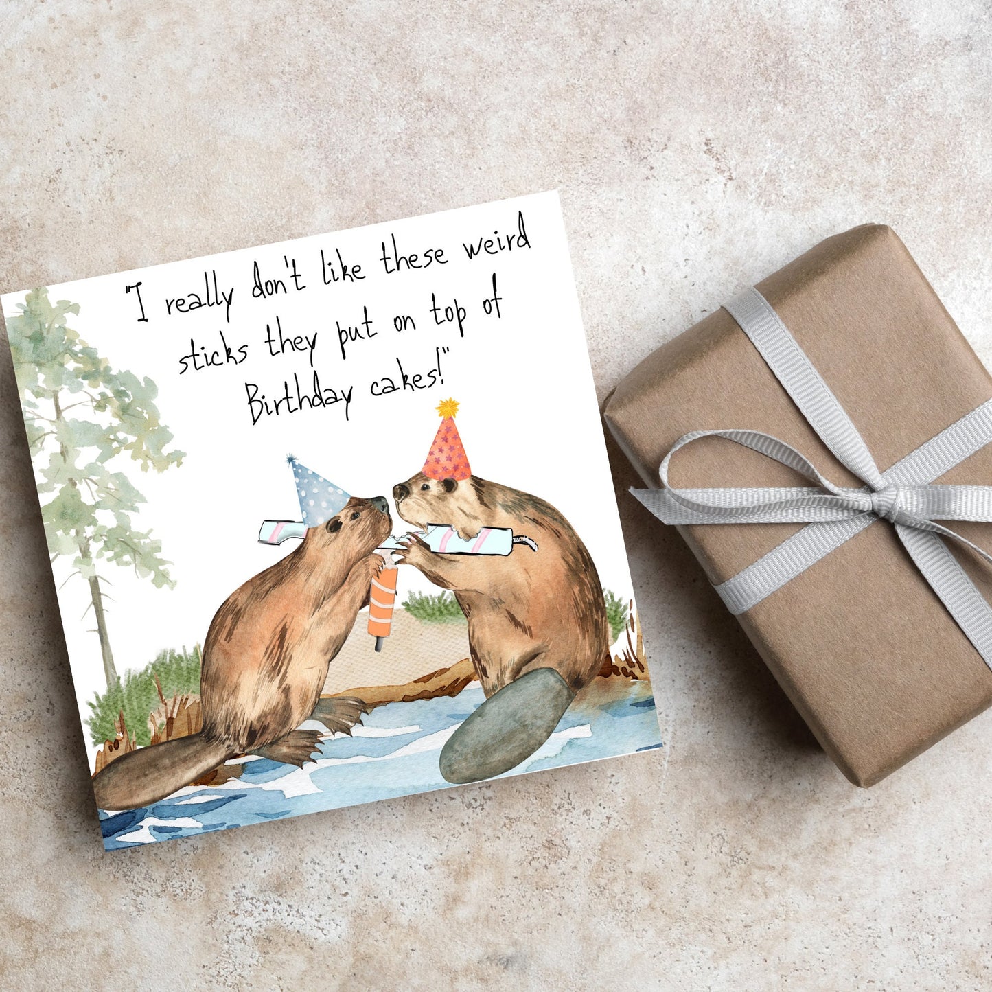 Image of a square birthday card with a watercolour image of 2 beavers with party hats on, chewing candles and handwritten style text above them that reads - i really don't like these weird sticks they put on top of birthday cakes!