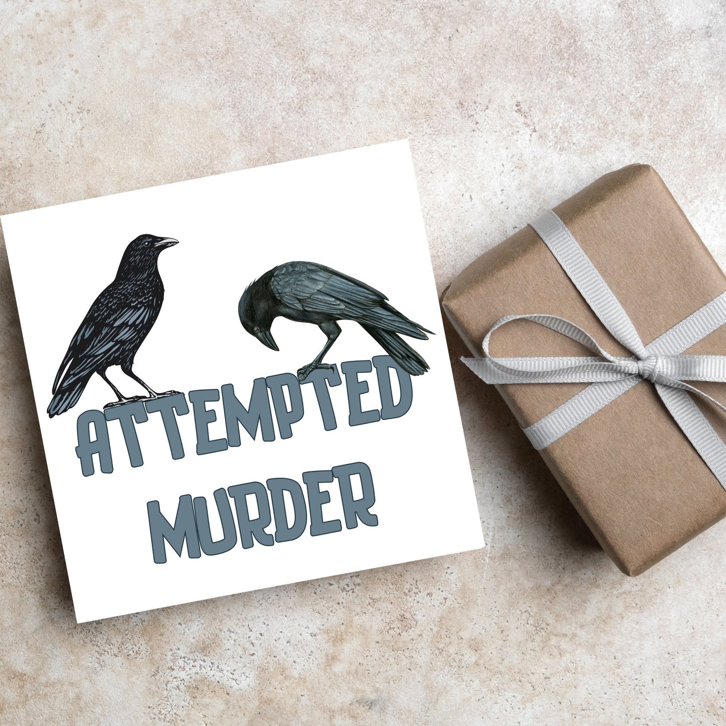 square card with the words attempted murder and 2 crows sitting on top of the text (a murder is the collective name 3 or more crows)