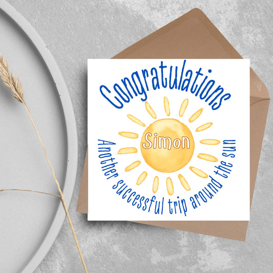 square birthday card with a watercolour image of a shining sun with a name in the middle of it in white text. Circled around the sun in blue text is written congratulations another successful trip around the sun