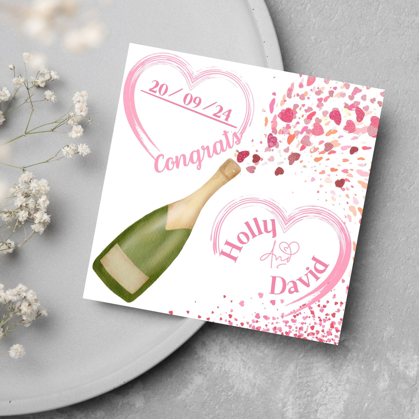 square wedding card with a bottle of champagne bursting with hearts in varying shades of pink. Above the bottle is a heart that holds the date and says contrats, and below is a repeated heart with the names of the couple inside