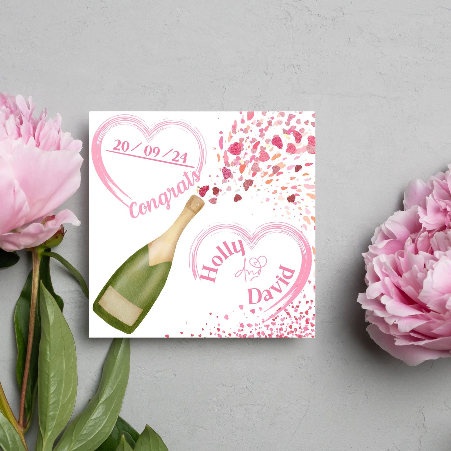 square wedding card with a bottle of champagne bursting with hearts in varying shades of pink. Above the bottle is a heart that holds the date and says contrats, and below is a repeated heart with the names of the couple inside