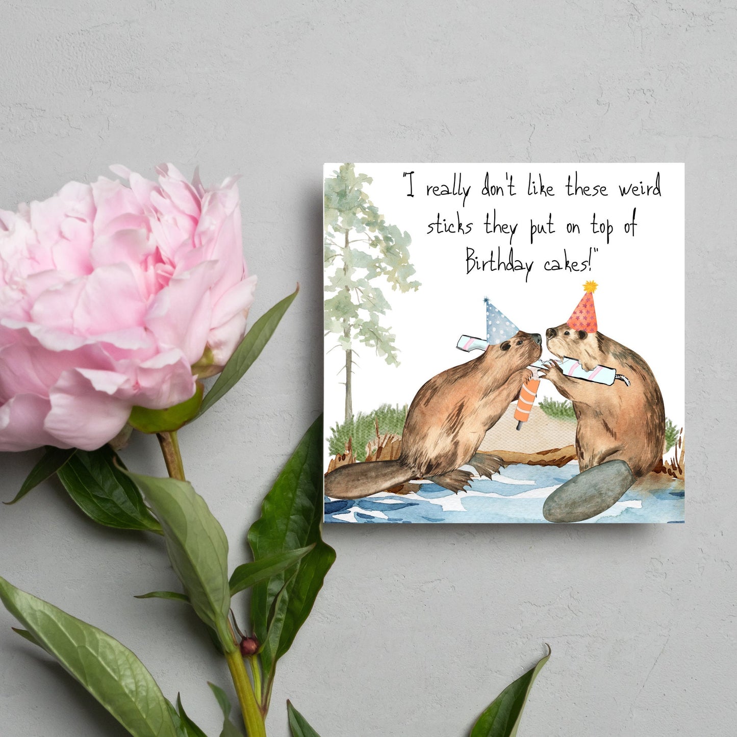 Image of a square birthday card with a watercolour image of 2 beavers with party hats on, chewing candles and handwritten style text above them that reads - i really don't like these weird sticks they put on top of birthday cakes!