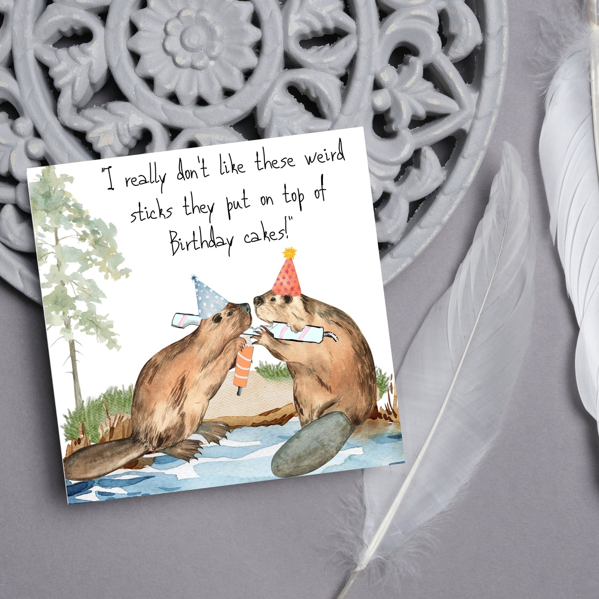 Image of a square birthday card with a watercolour image of 2 beavers with party hats on, chewing candles and handwritten style text  above them that reads - i really don't like these weird sticks they put on top of birthday cakes!