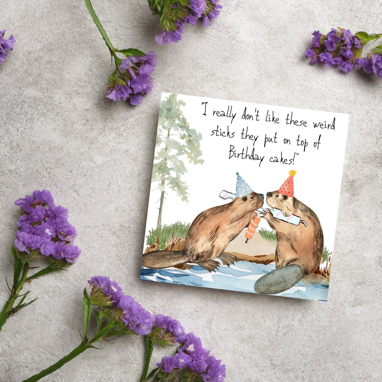 Image of a square birthday card with a watercolour image of 2 beavers with party hats on, chewing candles and handwritten style text above them that reads - i really don't like these weird sticks they put on top of birthday cakes!