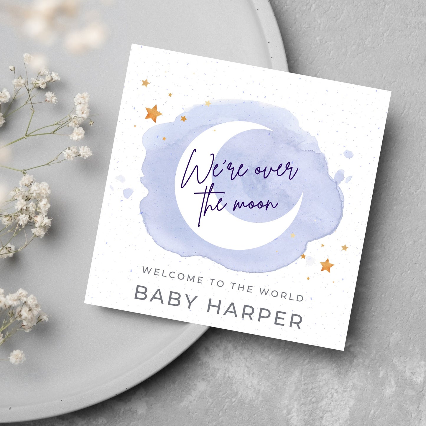 square card with a watercolour light blue background with a white crescent moon and yellow stars with navy blue hand script that reads - we're over the moon, and at the bottom in grey text welcome to the world baby harper