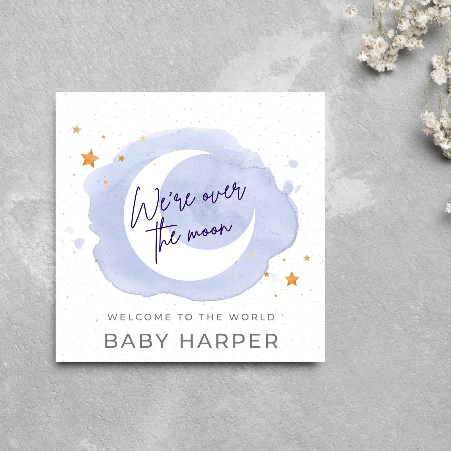 square card with a watercolour light blue background with a white crescent moon and yellow stars with navy blue hand script that reads - we're over the moon, and at the bottom in grey text welcome to the world baby harper