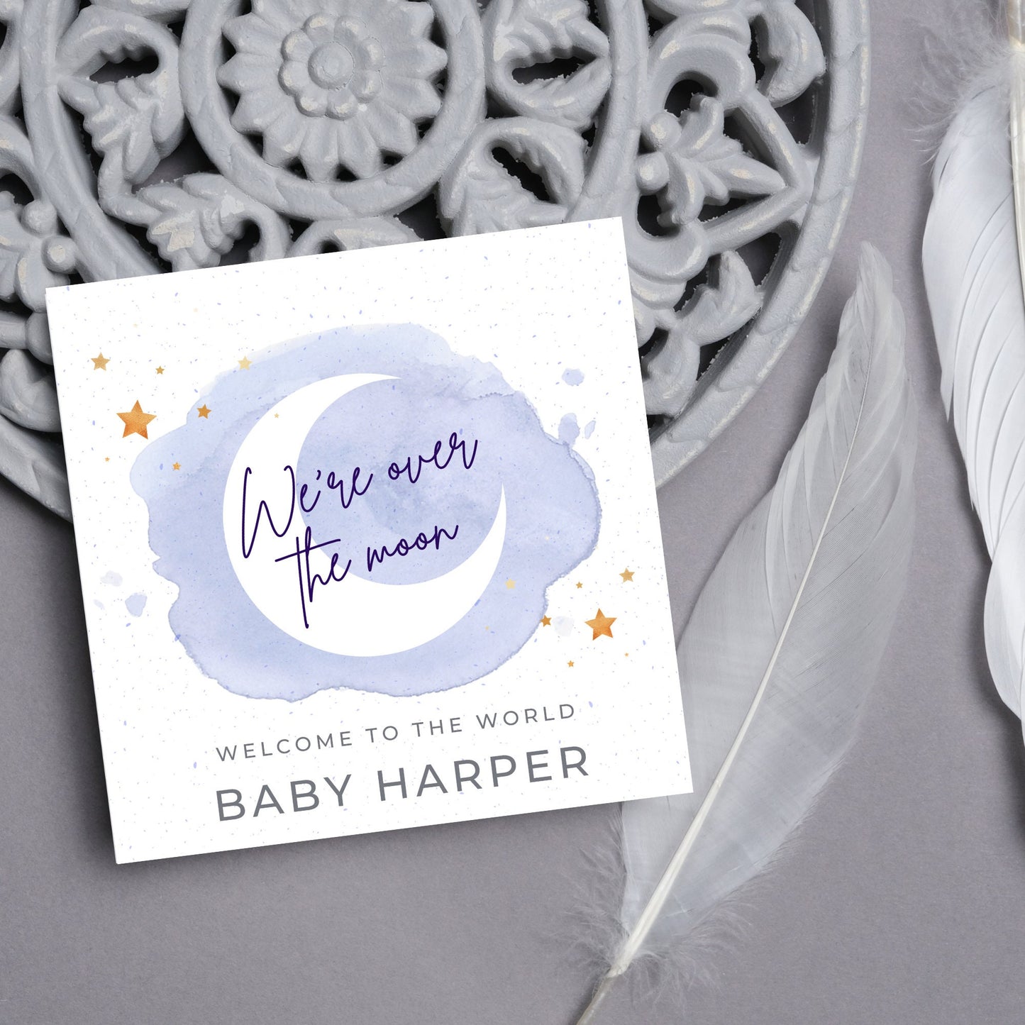 square card with a watercolour light blue background with a white crescent moon and yellow stars with navy blue hand script that reads - we're over the moon, and at the bottom in grey text welcome to the world baby harper